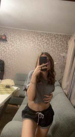 A woman with wavy brown hair takes a mirror selfie in a gray crop top and black shorts, standing in a living room with