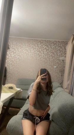 A woman with shoulder-length brown hair takes a mirror selfie while sitting on a gray couch, wearing a gray crop top