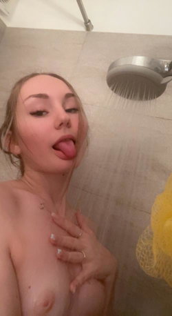 A woman with light hair sticks her tongue out in a shower, bare-chested, holding her chest with one hand while water