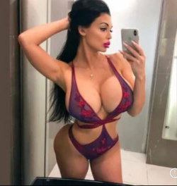 Brunette woman in purple lace lingerie takes a mirror selfie in a bathroom, showcasing a curvy figure with long dark