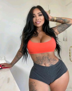 Brunette woman with long dark hair, curvy build, wearing orange crop top and gray shorts, posing indoors with visible