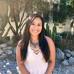A smiling woman with long dark hair wears a white lace-up top and statement necklace, standing outdoors near cacti and