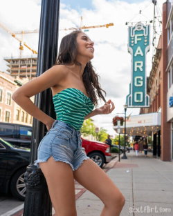A smiling woman with long dark hair wears a green striped top and denim shorts, leaning against a pole on a city