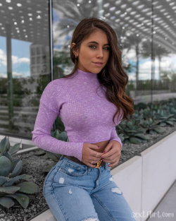 Brunette woman in a purple crop top and ripped jeans poses outdoors near succulents and glass walls.