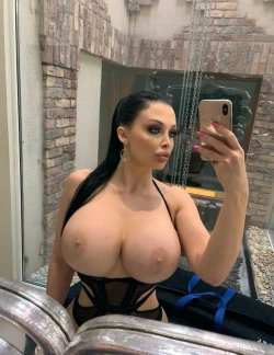 A woman with long dark hair takes a mirror selfie in a black lace top, showing bare breasts, in a brick-walled room.