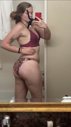 A woman with light brown hair in a bun takes a mirror selfie in a maroon bra and patterned bottoms, showing a curvy
