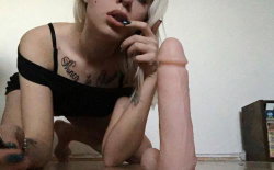 Blonde woman in black top with visible tattoos, kneeling on floor holding a large pink dildo, hand near mouth.