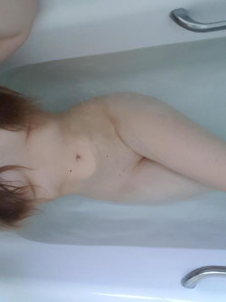 A person with reddish hair lies in a bathtub, partially submerged, showing bare shoulders and chest.