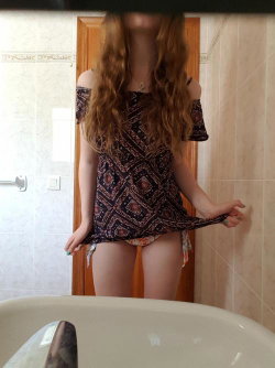 A woman with long curly hair stands in a bathroom, lifting a patterned off-shoulder top to reveal floral underwear.