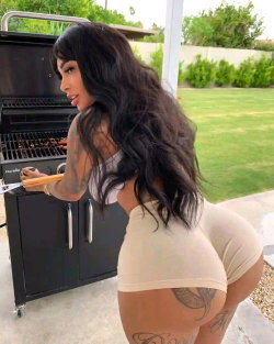 Brunette woman with long wavy hair and visible tattoos grilling outdoors, wearing a tight beige outfit.