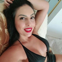 Brunette woman with red lipstick in a black top, lying on a plush surface, smiling at the camera.