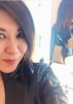 Woman with shoulder-length dark hair in a shiny black top, smiling slightly in a mirror selfie indoors.