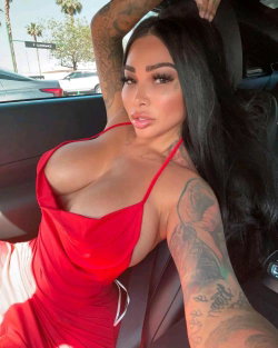 A woman with long dark hair and full tattoos on her arms poses in a red dress inside a car, looking at the camera.