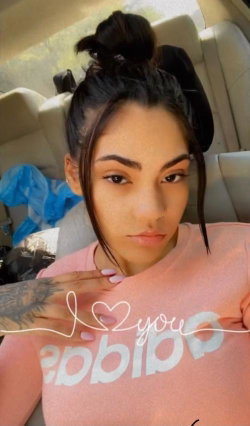 Brunette woman with tattoos on her arm, wearing a pink shirt, taking a selfie in a car.