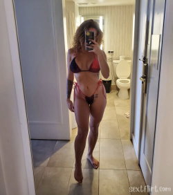 A woman with curly hair takes a mirror selfie in a red and black bikini, standing barefoot in a bathroom with tiled