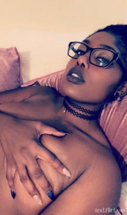 A Black woman with glasses and dark lipstick lies on a bed, wearing a choker, her hand resting over her bare chest.