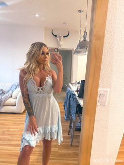 Blonde woman in a white lace dress takes a mirror selfie in a modern living room with wooden floors and pendant lights.
