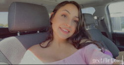 A woman with long dark hair smiles while sitting in a car, wearing a pink off-the-shoulder top.