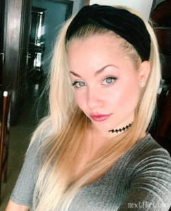 Blonde woman with long hair, wearing a black headband and choker, in a gray ribbed top, taking a selfie indoors.