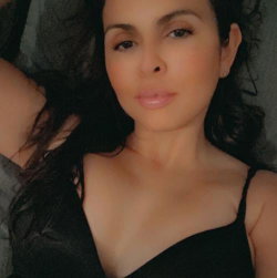A woman with dark curly hair lies on a gray surface, wearing a black spaghetti strap top, looking directly at the