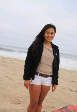 A smiling woman with long dark hair stands on a sandy beach, wearing a black jacket over a beige top and white shorts.
