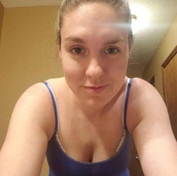 Woman with brown hair tied back, wearing a blue tank top, taking a close-up selfie indoors.