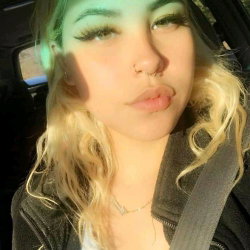 Blonde woman with wavy hair and nose ring taking a selfie in a car, wearing a dark jacket and delicate necklace.