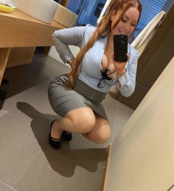 A curvy woman with long red hair takes a mirror selfie in a light blue blouse and gray skirt, posing with one hand on