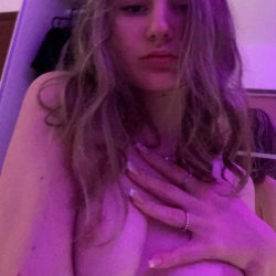 Blonde woman with wavy hair, bare shoulders, hand on chest, in dim purple lighting indoors.