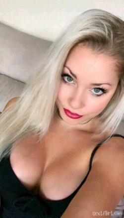 Blonde woman with long hair and red lipstick wearing a black top, posing closely on a couch in a selfie-style shot.