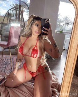 A woman with long blonde hair and tattoos takes a mirror selfie in a red heart-patterned bikini, sitting on a bed in a