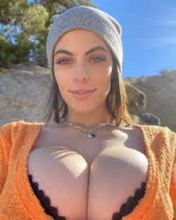 A woman with dark hair wearing a gray beanie and orange cardigan, standing outdoors in sunlight with bare breasts