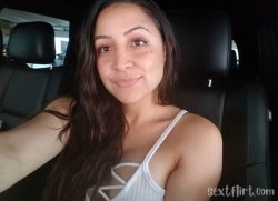 A woman with long dark hair smiles in a car selfie, wearing a white strappy top.