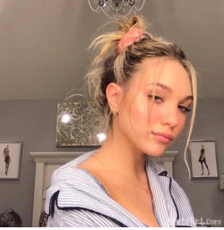 Blonde woman with hair in a messy bun, wearing a striped button-up shirt, posing indoors near framed artwork.