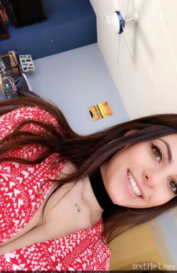 Brunette woman in a red heart-patterned top with a black choker, smiling in a selfie-style shot indoors.