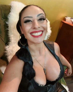 A smiling woman with dark hair in a braid, wearing a black leather top and large hoop earrings, posing indoors near a