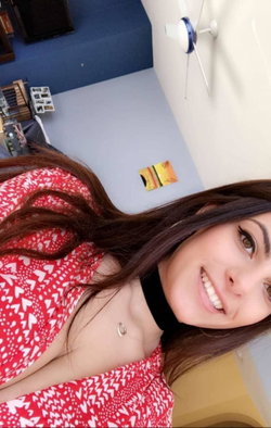Brunette woman in a red heart-patterned top with a black choker, smiling in a selfie-style photo indoors.