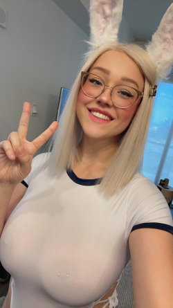 Blonde woman with glasses and bunny ears wears a tight white top, smiling while making a peace sign in a room with a