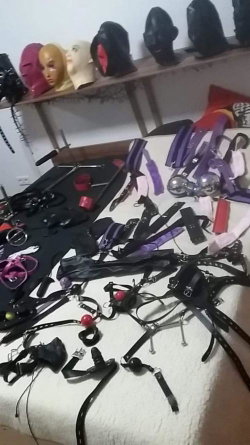 Bed covered with various BDSM gear including restraints, collars, and masks; mannequin heads on shelf in background.