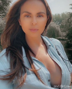 Brunette woman with long hair in a light blue unbuttoned shirt, standing outdoors near a flowering tree and water.