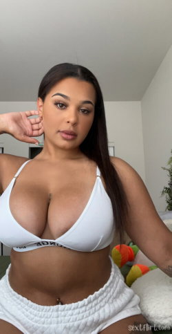 Curvy woman with long dark hair wearing a white bikini top and matching bottoms, posing indoors with one hand near her