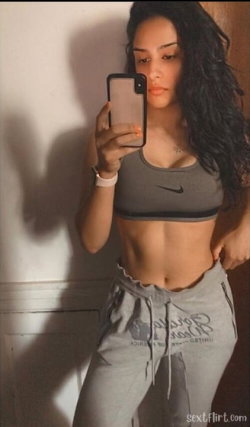 A woman with long dark hair takes a mirror selfie in a gray sports bra and sweatpants, showing toned abs against a