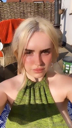 Blonde woman in a green halter top takes a selfie outdoors on a sunny patio with wicker furniture.