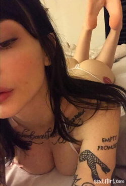 Brunette woman with visible tattoos on neck and arm, wearing a light pink top, lying down in a close-up selfie pose.