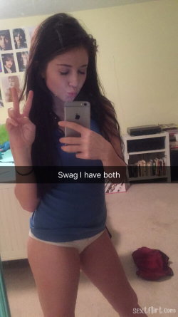Brunette woman in a blue crop top and white thong takes a mirror selfie, making a peace sign in a bedroom setting.