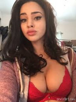 Brunette woman with long dark hair wearing a red lace bra and open hoodie, taking a selfie indoors.