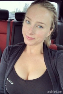 Blonde woman with blue eyes in a black top and cardigan, taking a selfie inside a car with red seats.