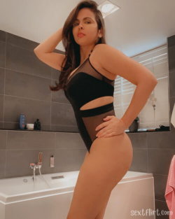 Curvy woman with long dark hair poses in a black mesh bodysuit in a modern bathroom with gray tiles and a white tub.