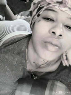 A Black woman with a floral headwrap and neck tattoo rests her head on a striped pillow, looking at the camera in a