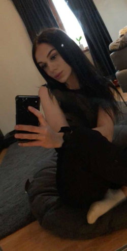 A woman with long dark hair sits on a beanbag, holding her phone for a selfie, wearing black clothing in a cozy room.